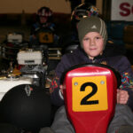 Ein Kind mit Hut sitzt in einem Go-Kart mit der Nummer 2, während im Hintergrund andere Rennfahrer und Karts zu sehen sind.