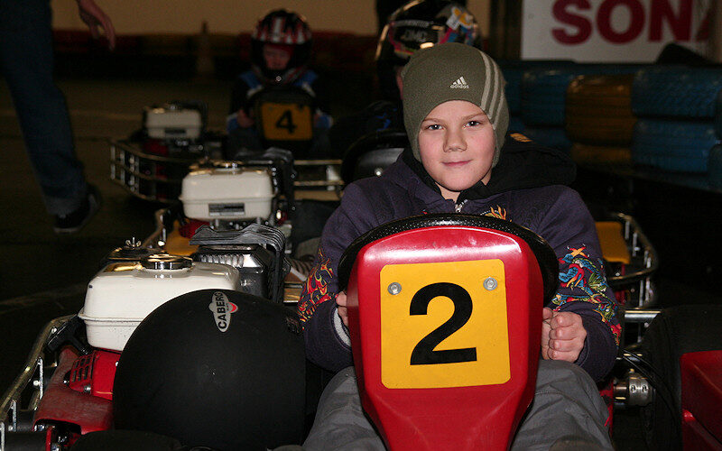 Ein Kind mit Hut sitzt in einem Go-Kart mit der Nummer 2, während im Hintergrund andere Rennfahrer und Karts zu sehen sind.