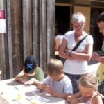 Erwachsene stehen um eine Gruppe von Kindern herum, die an einem sonnigen Tag an einem Tisch sitzen und sich mit einer künstlerischen oder handwerklichen Tätigkeit im Freien beschäftigen.