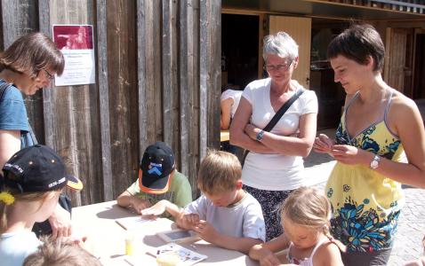 Erwachsene stehen um eine Gruppe von Kindern herum, die an einem sonnigen Tag an einem Tisch sitzen und sich mit einer künstlerischen oder handwerklichen Tätigkeit im Freien beschäftigen.