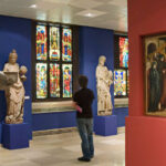 Eine Person steht in einer Museumsgalerie mit Buntglasfenstern, Skulpturen auf Sockeln und einem religiösen Gemälde an einer roten Wand.