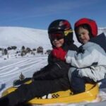 Zwei Kinder mit Helmen und Winterjacken sitzen zusammen auf einem gelben Schlitten auf einem verschneiten Hügel, mit einer verschneiten Landschaft im Hintergrund.