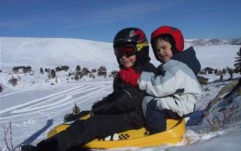 Zwei Kinder mit Helmen und Winterjacken sitzen zusammen auf einem gelben Schlitten auf einem verschneiten Hügel, mit einer verschneiten Landschaft im Hintergrund.