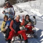 Eine Gruppe von sechs Personen, darunter auch Kinder, sitzt in einer verschneiten Umgebung auf Schlitten zusammen und winkt der Kamera zu.