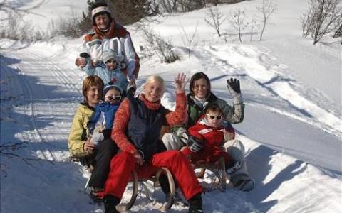 Eine Gruppe von sechs Personen, darunter auch Kinder, sitzt in einer verschneiten Umgebung auf Schlitten zusammen und winkt der Kamera zu.