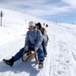 Zwei Personen in Winterkleidung schlittern unter teilweise bewölktem Himmel einen verschneiten Hügel hinunter.