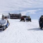 Eine Gruppe von Menschen fährt mit dem Schlitten einen verschneiten Hügel hinunter, mit einem Gebäude im Hintergrund unter einem bewölkten Himmel.