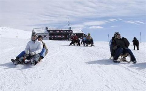 Eine Gruppe von Menschen fährt mit dem Schlitten einen verschneiten Hügel hinunter, mit einem Gebäude im Hintergrund unter einem bewölkten Himmel.