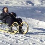 Ein winterlich gekleidetes Kind fährt auf einem Holzschlitten einen verschneiten Hügel hinunter.