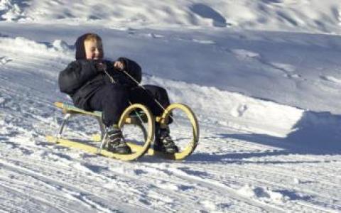 Ein winterlich gekleidetes Kind fährt auf einem Holzschlitten einen verschneiten Hügel hinunter.