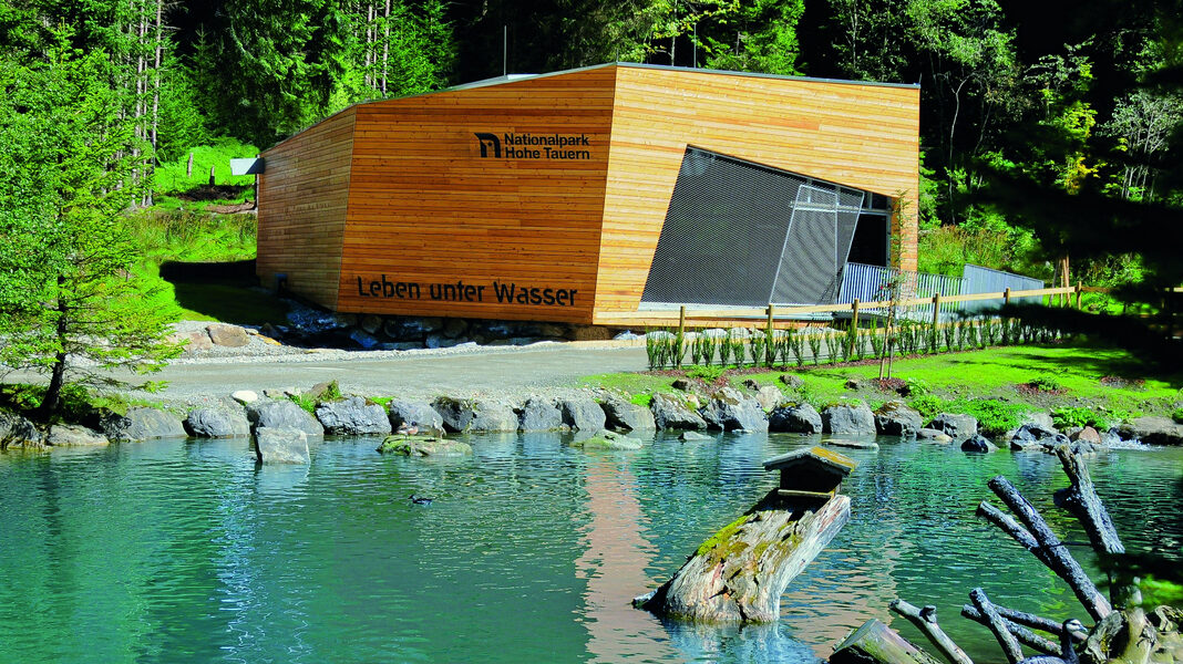 Ein modernes Holzgebäude mit der Aufschrift "Leben unter Wasser" steht an einem klaren, grünlichen Teich, umgeben von Felsen, Baumstämmen und dichtem Wald.