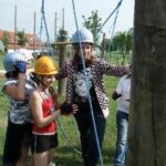 Vier Personen mit Helmen und Sicherheitsausrüstung stehen im Freien in einem Hochseilgarten und interagieren mit Seilen und Holzpfosten.