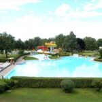 Außenschwimmbad mit klarem, blauem Wasser, umgeben von grünen Hecken und Bäumen, mit Wasserrutsche und freiem Himmel darüber.