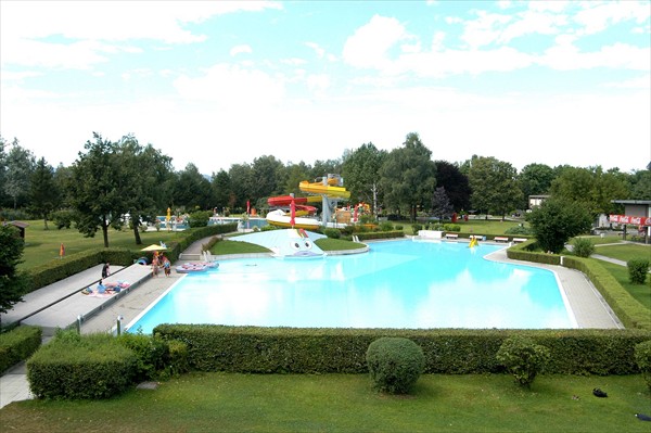 Außenschwimmbad mit klarem, blauem Wasser, umgeben von grünen Hecken und Bäumen, mit Wasserrutsche und freiem Himmel darüber.