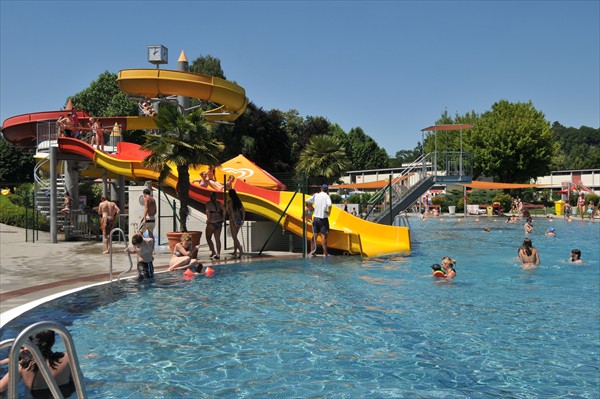 Menschen genießen einen sonnigen Tag in einem Freibad mit einer gelben und orangefarbenen Wasserrutsche. Einige schwimmen, während andere darauf warten, dass sie mit der Rutsche an der Reihe sind. Bäume und Sitzgelegenheiten am Beckenrand sind zu sehen.