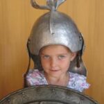 Ein junges Mädchen, das einen Metallhelm trägt und ein verziertes Metallschild hält, steht vor einem schlichten Hintergrund.