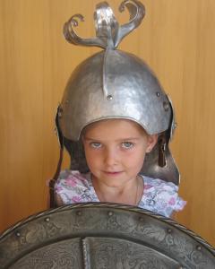 Ein junges Mädchen, das einen Metallhelm trägt und ein verziertes Metallschild hält, steht vor einem schlichten Hintergrund.