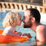 Ein Erwachsener und ein Kind berühren sich lächelnd in einem Schwimmbecken, wobei das Kind in einem orangefarbenen aufblasbaren Ring sitzt.