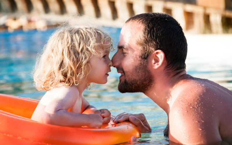 Ein Erwachsener und ein Kind berühren sich lächelnd in einem Schwimmbecken, wobei das Kind in einem orangefarbenen aufblasbaren Ring sitzt.