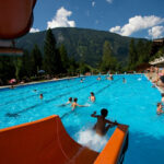 Freibad mit vielen Menschen, die das Wasser genießen; eine Wasserrutsche im Vordergrund, Berge, Bäume und ein blauer Himmel im Hintergrund.