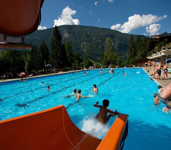 Freibad mit vielen Menschen, die das Wasser genießen; eine Wasserrutsche im Vordergrund, Berge, Bäume und ein blauer Himmel im Hintergrund.