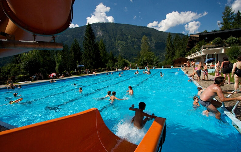 Freibad mit vielen Menschen, die das Wasser genießen; eine Wasserrutsche im Vordergrund, Berge, Bäume und ein blauer Himmel im Hintergrund.