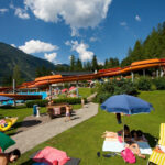 An einem sonnigen Tag entspannt man sich auf Liegestühlen und Handtüchern unter Sonnenschirmen in der Nähe einer Wasserrutsche im Freien, umgeben von Gras, Bäumen und Bergen.