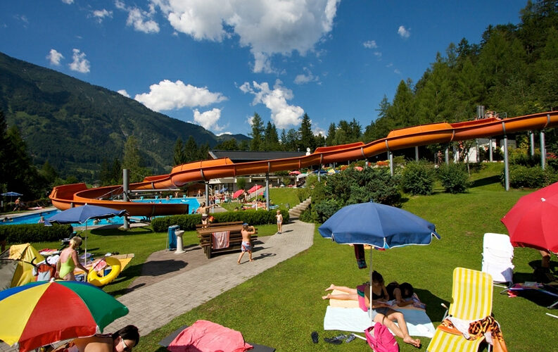 An einem sonnigen Tag entspannt man sich auf Liegestühlen und Handtüchern unter Sonnenschirmen in der Nähe einer Wasserrutsche im Freien, umgeben von Gras, Bäumen und Bergen.