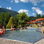 Außenpool mit Kindern und Erwachsenen im flachen Wasser, umgeben von Grün, Rutschen und Bergen unter einem teilweise bewölkten Himmel.