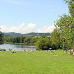 Menschen entspannen sich auf einer Wiese neben einem See mit Bäumen und Hügeln im Hintergrund unter einem teilweise bewölkten Himmel.