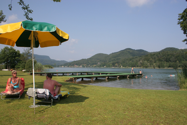 Menschen entspannen sich auf Liegestühlen unter einem Sonnenschirm in der Nähe eines Stegs an einem See, mit Schwimmern im Wasser und grünen Hügeln im Hintergrund.