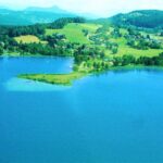 Luftaufnahme eines blauen Sees mit einer kleinen Halbinsel, umgeben von grünen Feldern, Bäumen und entfernten Hügeln unter einem teilweise bewölkten Himmel.