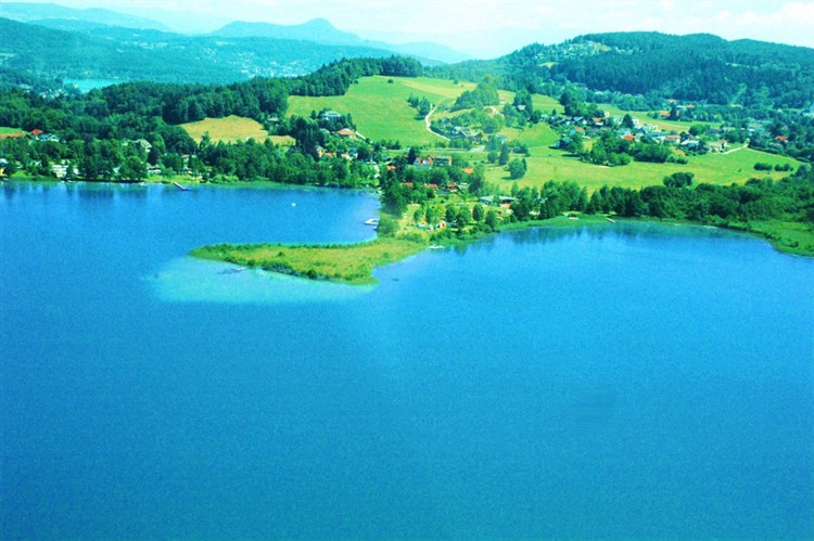 Luftaufnahme eines blauen Sees mit einer kleinen Halbinsel, umgeben von grünen Feldern, Bäumen und entfernten Hügeln unter einem teilweise bewölkten Himmel.
