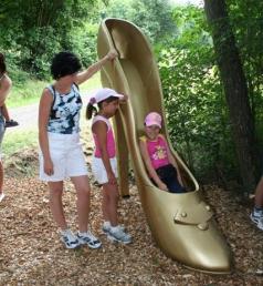 Eine Frau und zwei Kinder stehen im Freien neben einer riesigen goldenen Schuhskulptur mit hohen Absätzen; ein Kind sitzt im Inneren des Schuhs.