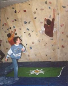 Drei Kinder klettern und spielen an einer Indoor-Kletterwand, unter ihnen liegt eine grüne Matte auf dem gepolsterten Boden.