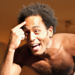 Eine hemdsärmelige Person mit lockigem Haar deutet auf ihre Stirn und lächelt. Sie wirkt ausdrucksstark und lebendig in einer warm beleuchteten Umgebung.