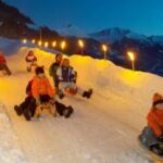 Eine Gruppe von Menschen schlittelt in der Dämmerung eine verschneite Piste hinunter, umgeben von Schneewänden und brennenden Fackeln, mit den Bergen im Hintergrund.