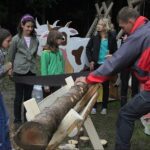 Ein Mann sägt mit einer Handsäge einen großen Baumstamm, während vier Mädchen in der Nähe von hölzernen Tierfiguren und einer tragbaren Toilette zusehen.