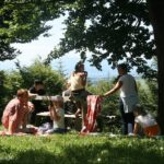 Eine Gruppe von Menschen, darunter Erwachsene und Kinder, macht ein Picknick auf einer Wiese unter Bäumen mit einem hölzernen Picknicktisch und einem malerischen Hintergrund.
