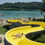 Eine Person rutscht eine leuchtend gelbe Wasserrutsche in der Nähe eines Sees hinunter, während im Hintergrund an einem sonnigen Tag Menschen sonnenbaden, schwimmen und sich entspannen.