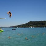 Eine Person springt von einem Sprungbrett in einen See, in dem mehrere Personen schwimmen und ein grünes Schlauchboot im Wasser liegt. Im Hintergrund sind bewaldete Hügel und Segelboote zu sehen.