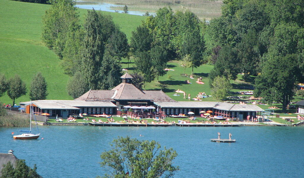 Ein Erholungsgebiet am See mit einem großen Gebäude, Sonnenanbetern auf Rasenflächen und Menschen, die schwimmen, paddeln und sich am Wasser entspannen, umgeben von Bäumen und Hügeln.