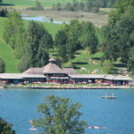 Ein Erholungsgebiet am See mit einem großen Gebäude, Sonnenanbetern auf Rasenflächen und Menschen, die schwimmen, paddeln und sich am Wasser entspannen, umgeben von Bäumen und Hügeln.