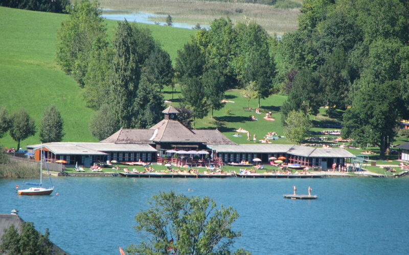 Ein Erholungsgebiet am See mit einem großen Gebäude, Sonnenanbetern auf Rasenflächen und Menschen, die schwimmen, paddeln und sich am Wasser entspannen, umgeben von Bäumen und Hügeln.