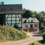 Zwei traditionelle Fachwerkhäuser mit weißen Wänden und dunklen Holzbalken sind an einem sonnigen Tag entlang eines Kiesweges von Grün umgeben.