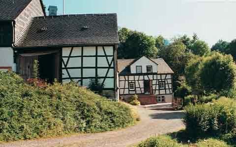 Zwei traditionelle Fachwerkhäuser mit weißen Wänden und dunklen Holzbalken sind an einem sonnigen Tag entlang eines Kiesweges von Grün umgeben.