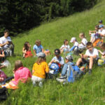 Eine Gruppe von Kindern und Erwachsenen sitzt auf einer grasbewachsenen Anhöhe und macht eine Pause oder ein Picknick, mit Rucksäcken um sie herum und Bäumen im Hintergrund.