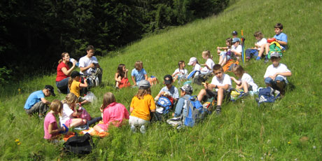 Eine Gruppe von Kindern und Erwachsenen sitzt auf einer grasbewachsenen Anhöhe und macht eine Pause oder ein Picknick, mit Rucksäcken um sie herum und Bäumen im Hintergrund.