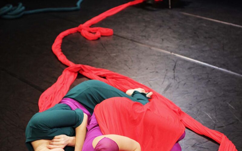 Zwei Darsteller liegen in rotem Stoff verschlungen auf der Bühne, während ein Musiker im Hintergrund Cello spielt.