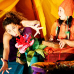 Zwei Frauen in farbenfrohen Kostümen treten auf; die eine streckt ausdrucksvoll eine große Blume in die Kamera, die andere sitzt daneben und hält eine Holzkiste, die mit einem gelben Zelt unterlegt ist.